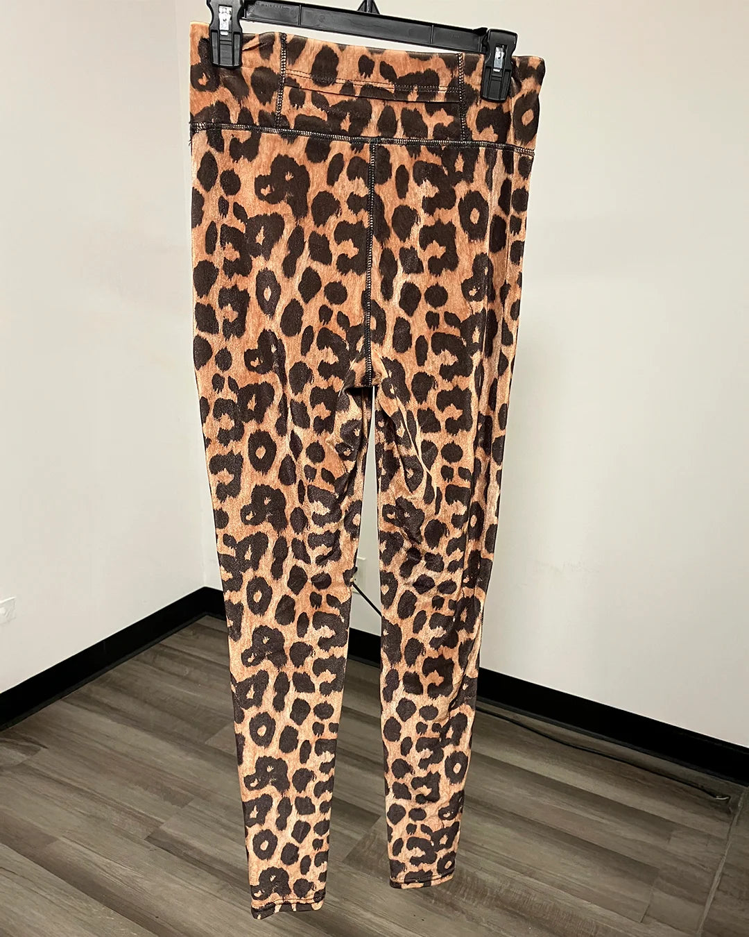 Leopard print leggings with a front flap-pocket hanging on a black hanger against a white wall and wood-look floor.