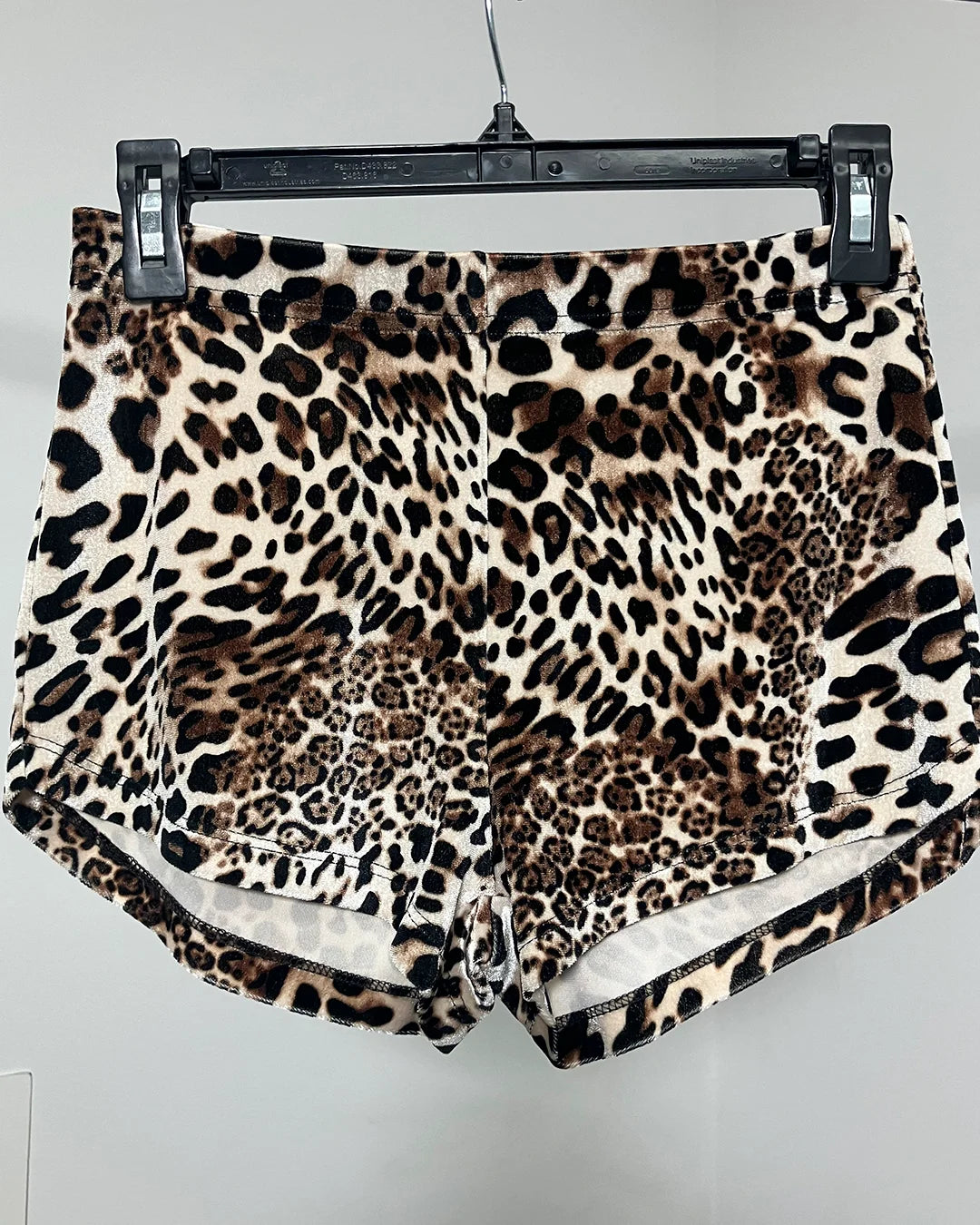 Leopard print shorts in a plush velvet-like fabric, hanging from a black plastic hanger against a light gray wall.