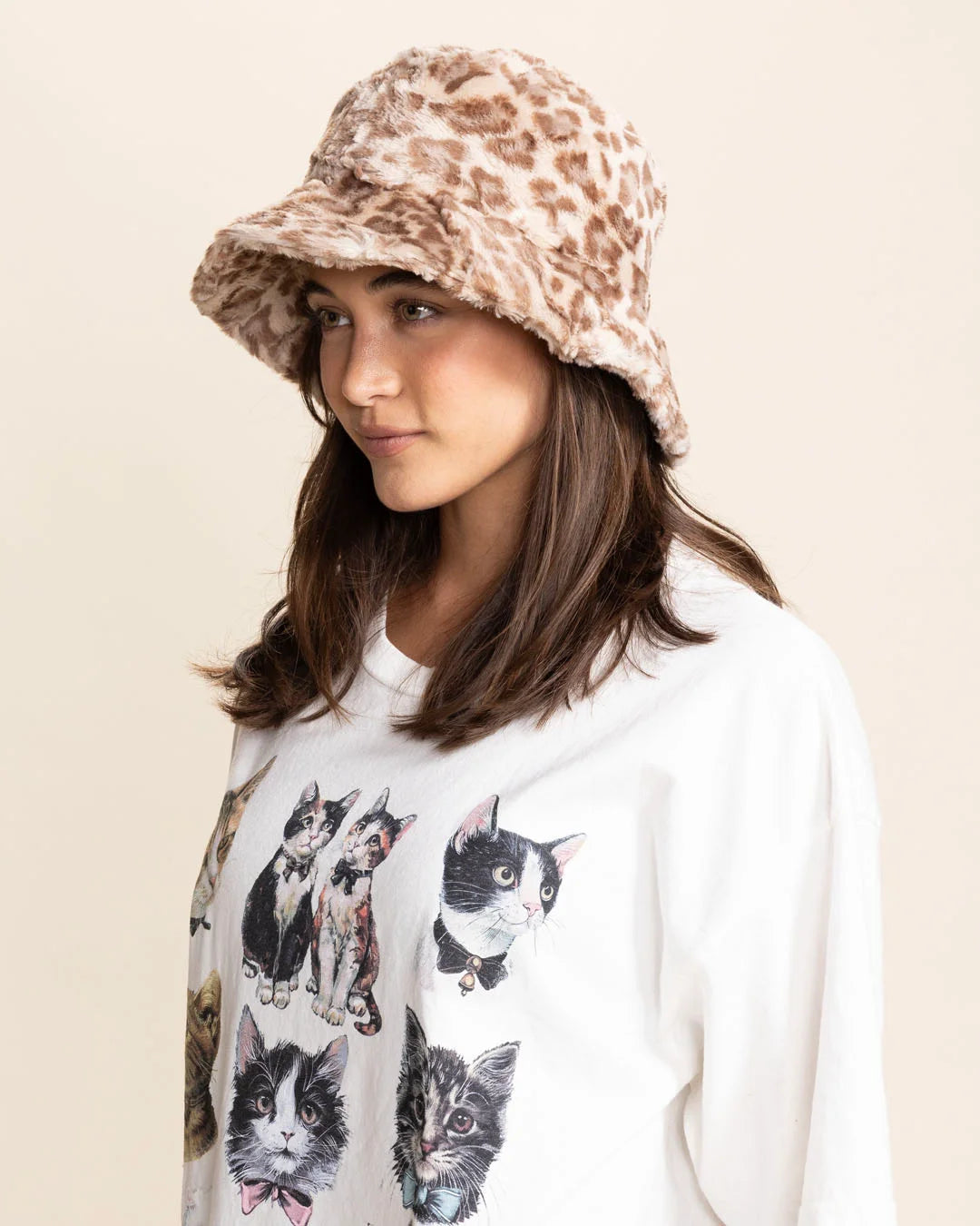 Woman wearing a tan leopard bucket hat and a white t-shirt with cat prints on a beige background