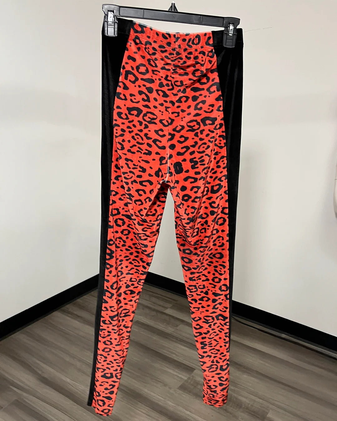 Bright orange and black leopard print leggings hanging from a black hanger against a white wall and wood-look floor.