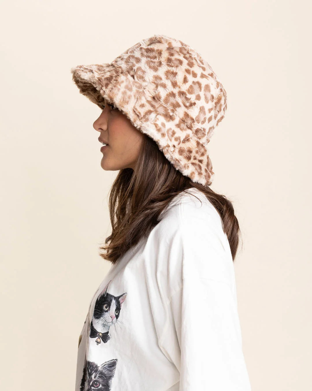 Person wearing a patterned tan leopard print fuzzy bucket hat and a white shirt with cat designs on a beige background