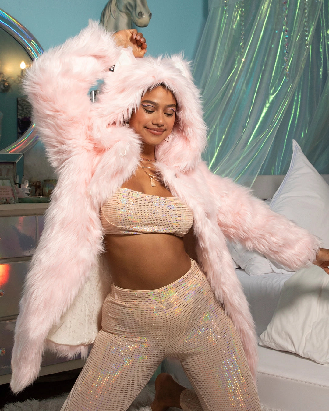 A Woman in a pink fake fur coat and gold sequin outfit in a room with decorative elements. She has her arm up and is smiling in a blissful state while wearing SpiritHoods 100% Vegan PETA-approved Flamingo Wolf Faux Fur Coat