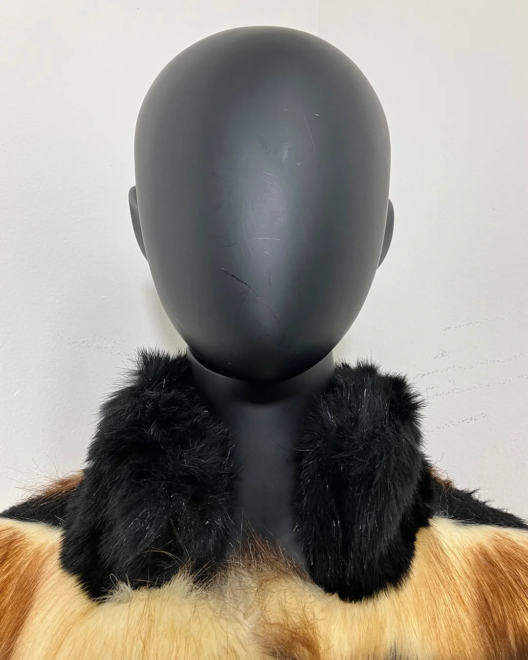 Mannequin head wearing a black faux fur collar with a multicolored garment underneath on a light gray background.