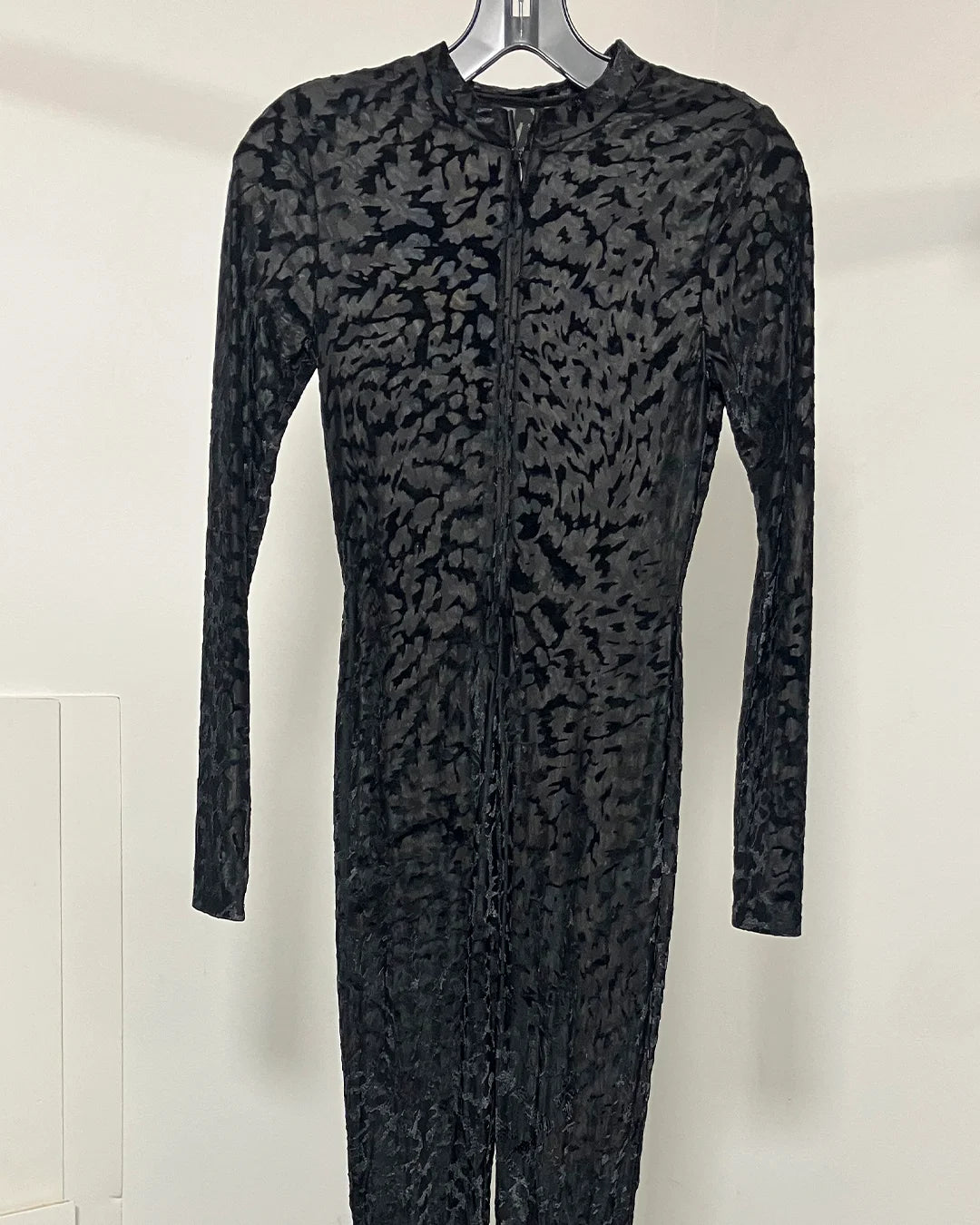 A long-sleeve black textured zip-front dress or jumpsuit displayed on a hanger against a plain light wall.