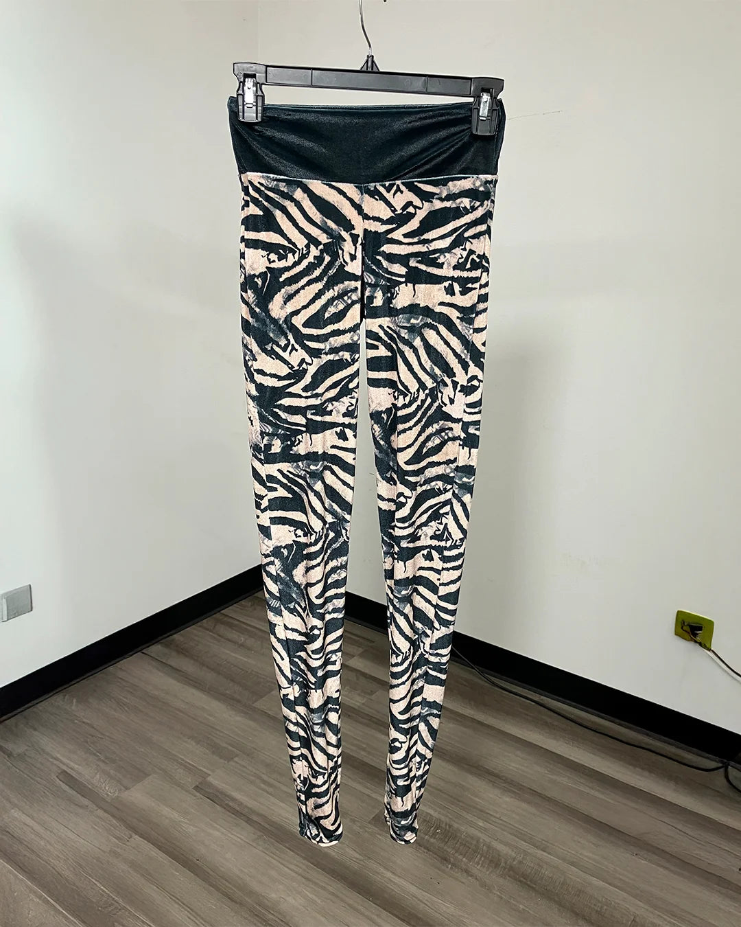 Zebra-print leggings hanging on a black hanger against a white wall with wood-look flooring.