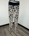 A pair of high-waisted leggings with a black and beige giraffe print pattern, hanging on a black hanger against a white wall and gray wood floor.