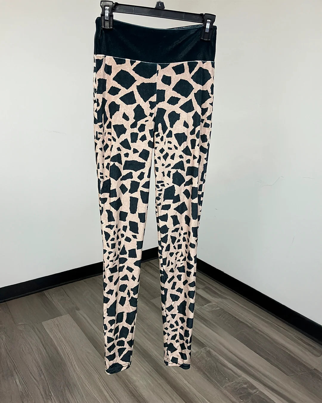 A pair of high-waisted leggings with a black and beige giraffe print pattern, hanging on a black hanger against a white wall and gray wood floor.