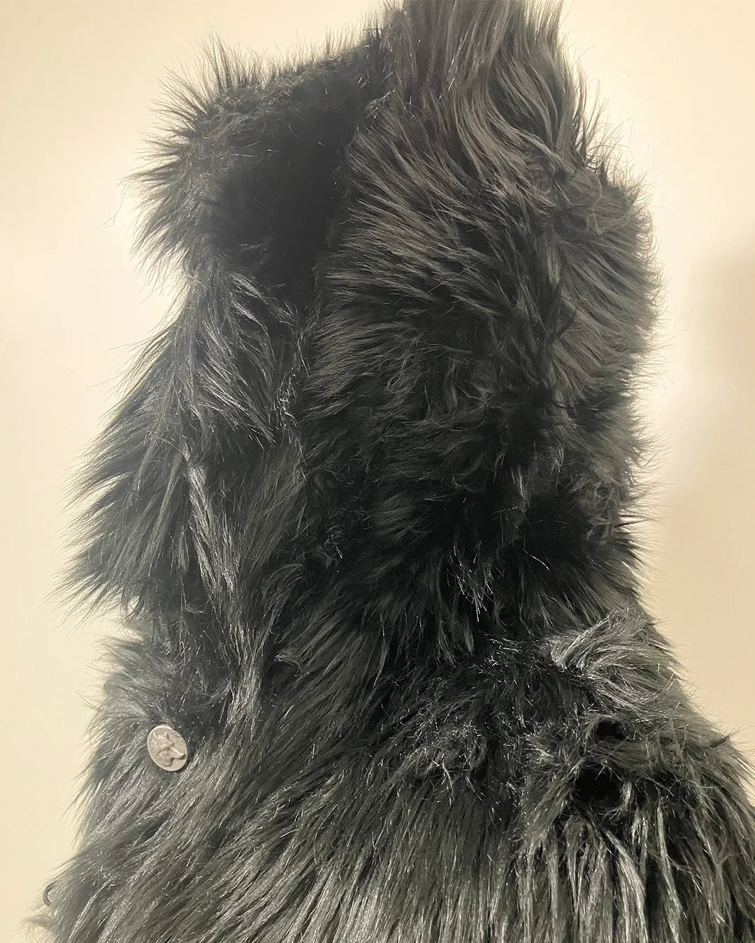 Close-up of a textured, black-colored fake fur with a wolf face button on a beige background. SpiritHoods XXS Sizing Black Wolf Online Sample Sale Style.