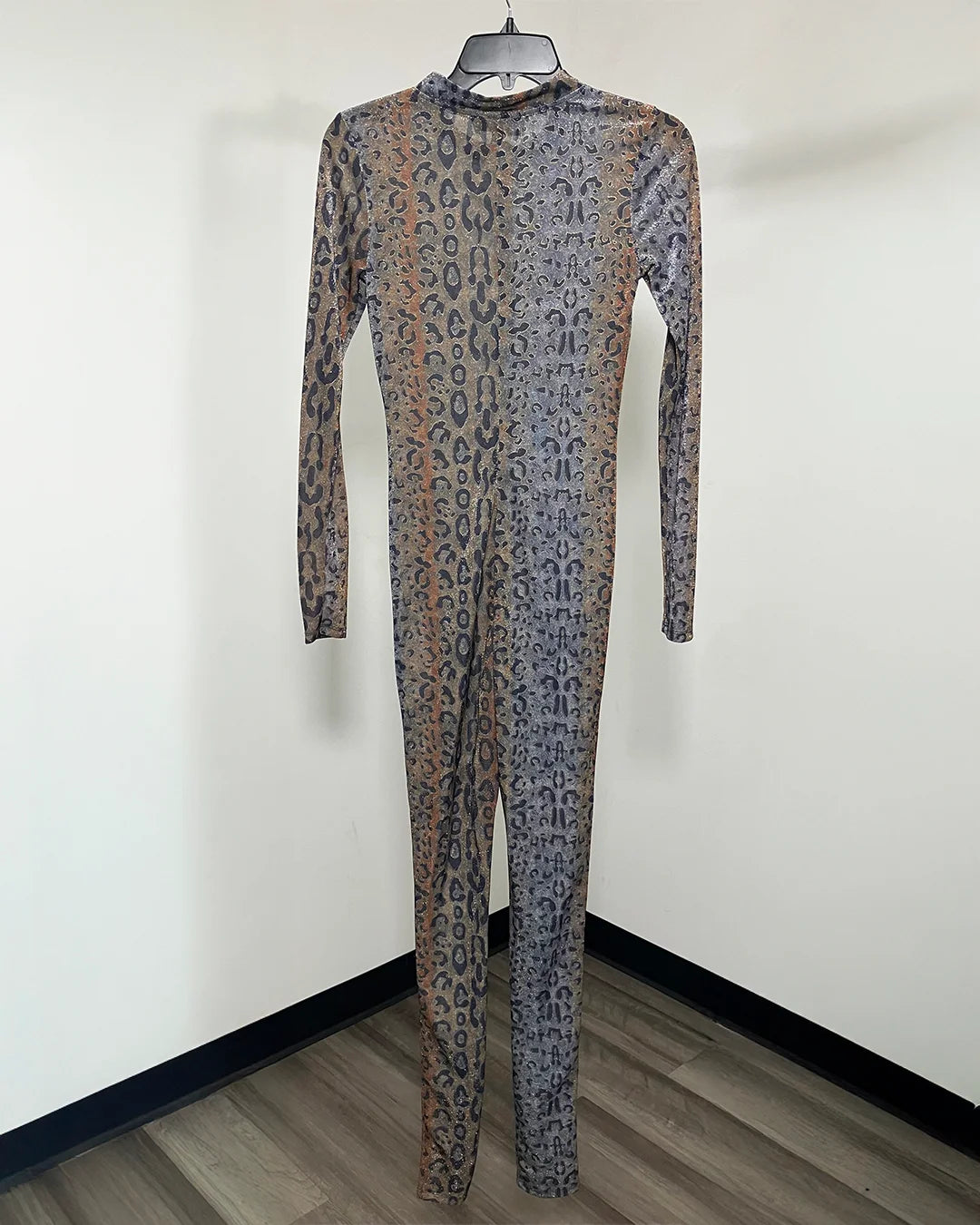 A long-sleeve full-body one-piece jumpsuit with an abstract leopard-like pattern in muted brown, gray, and black, hanging on a black hanger against a plain off-white wall.