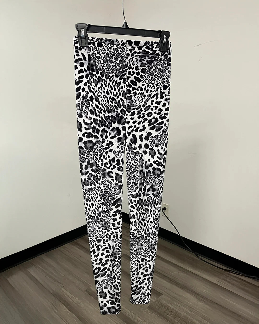 Black and white leopard print leggings hanging on a black hanger against an off-white wall with a wood floor.
