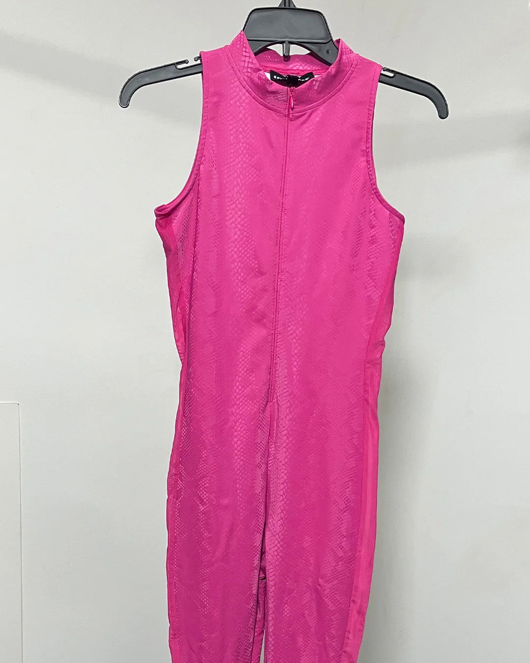 A bright pink snakeskin pattern sleeveless zip-front jumpsuit or bodysuit hanging on a black hanger against a plain light gray wall.