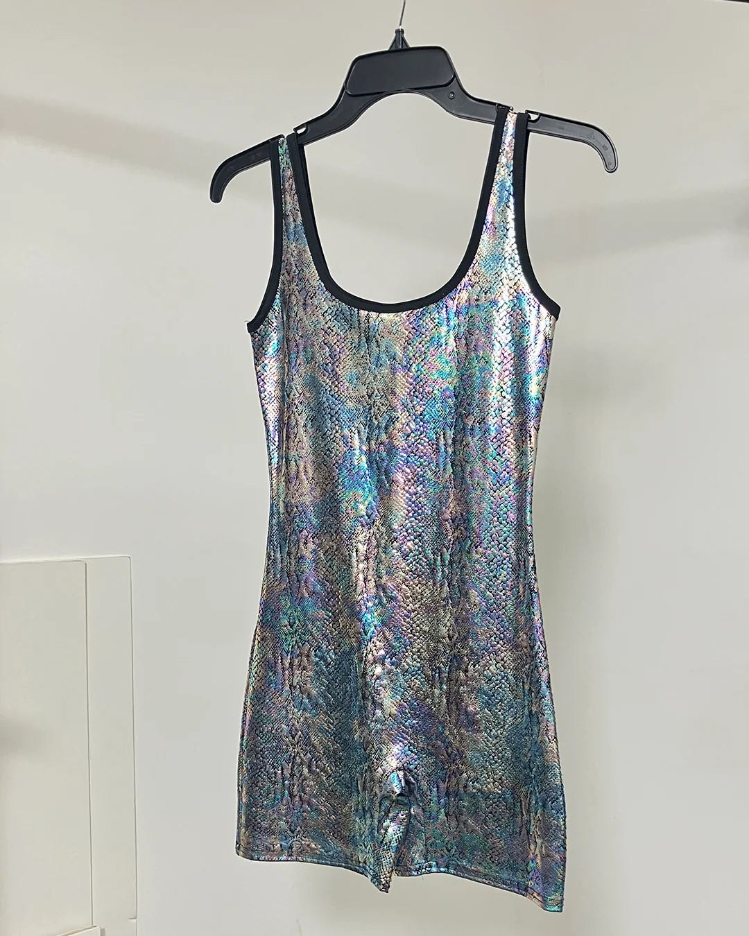 A sleeveless iridescent snakeskin-patterned dress with black trim, hanging on a black hanger against a plain white wall.