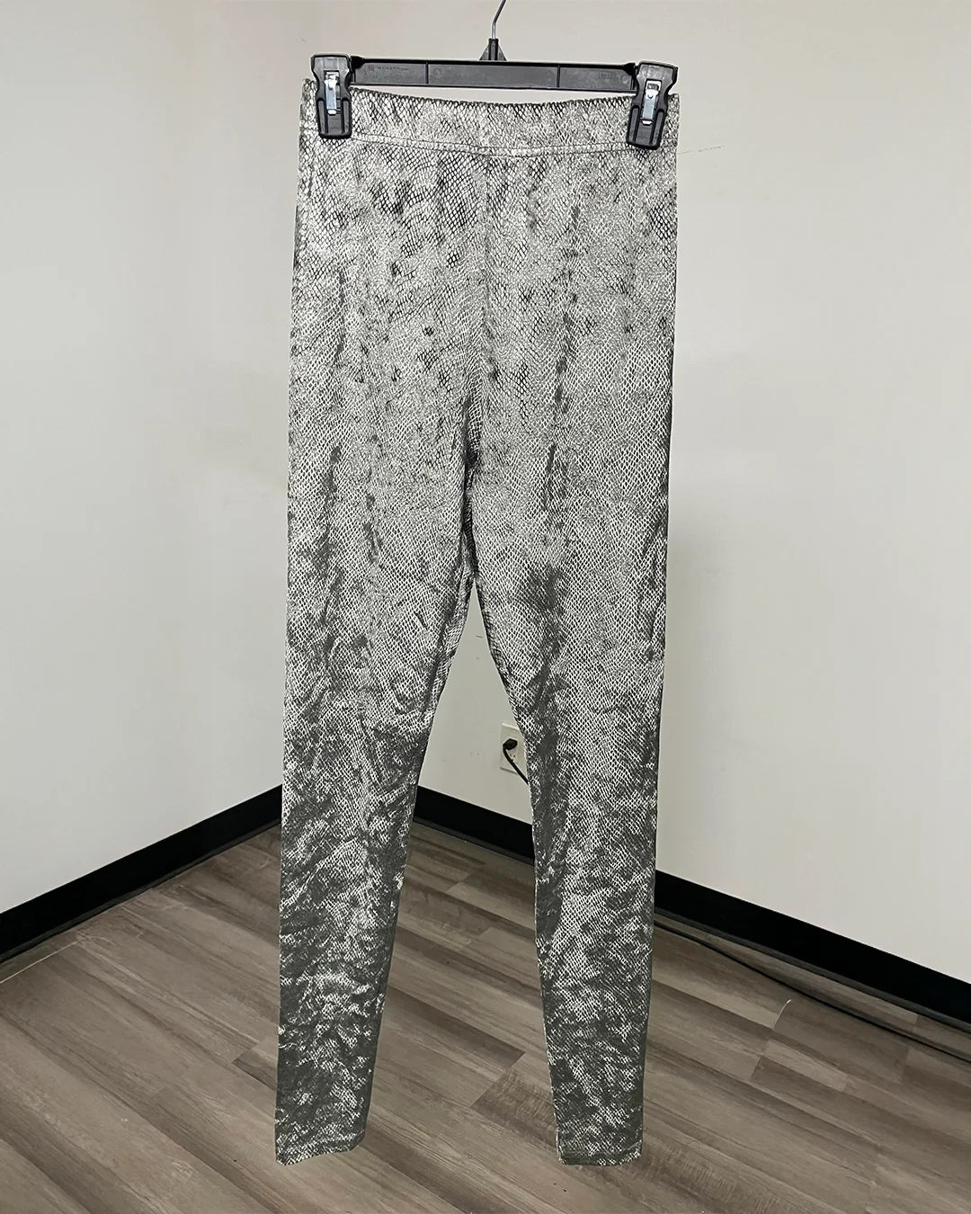 Greenish gray and black snakeskin print leggings hanging on a black hanger against a plain white wall with a wood floor.