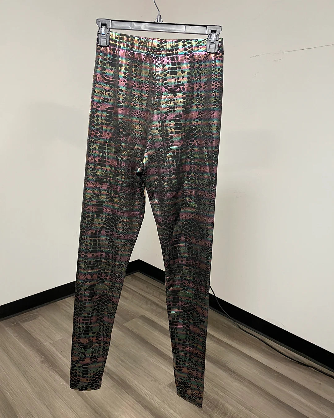 Metallic snakeskin print leggings hanging on a clear plastic hanger against a plain off-white wall with a wood-look floor.