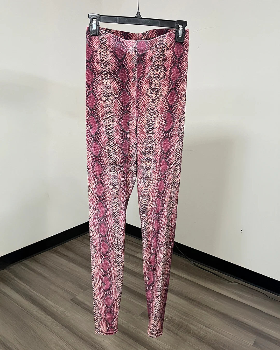 Pink and red snakeskin print leggings hanging on a black hanger against a plain off-white wall.