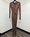 A long-sleeve, full-length leopard print jumpsuit hanging on a black hanger against a plain white wall with a wood-look floor.