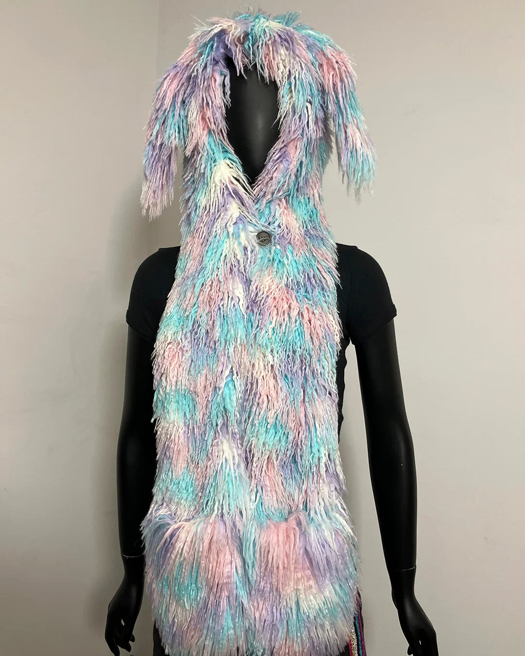 A long, shaggy faux-fur hood with a bunny ears, shown on a black mannequin. The vest has a pastel tie-dye pattern of pink, purple, blue, and white, with a small dark button at the neckline.