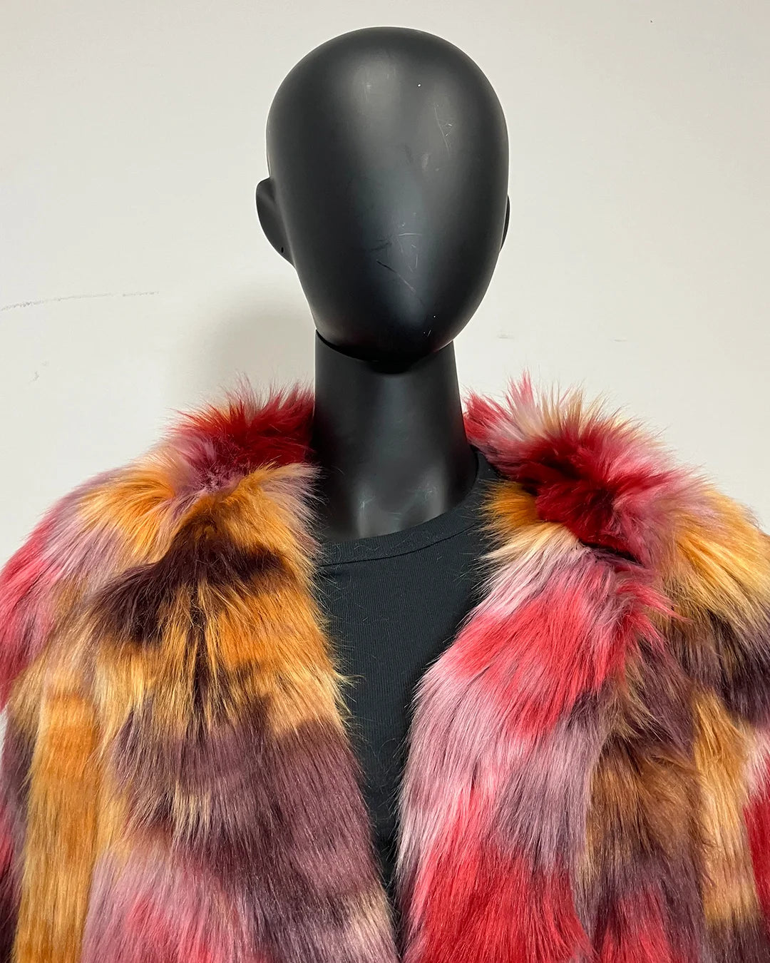 A multicolor faux fur jacket with red, orange, yellow, and purple tones, displayed on a black mannequin torso against a plain off-white wall.