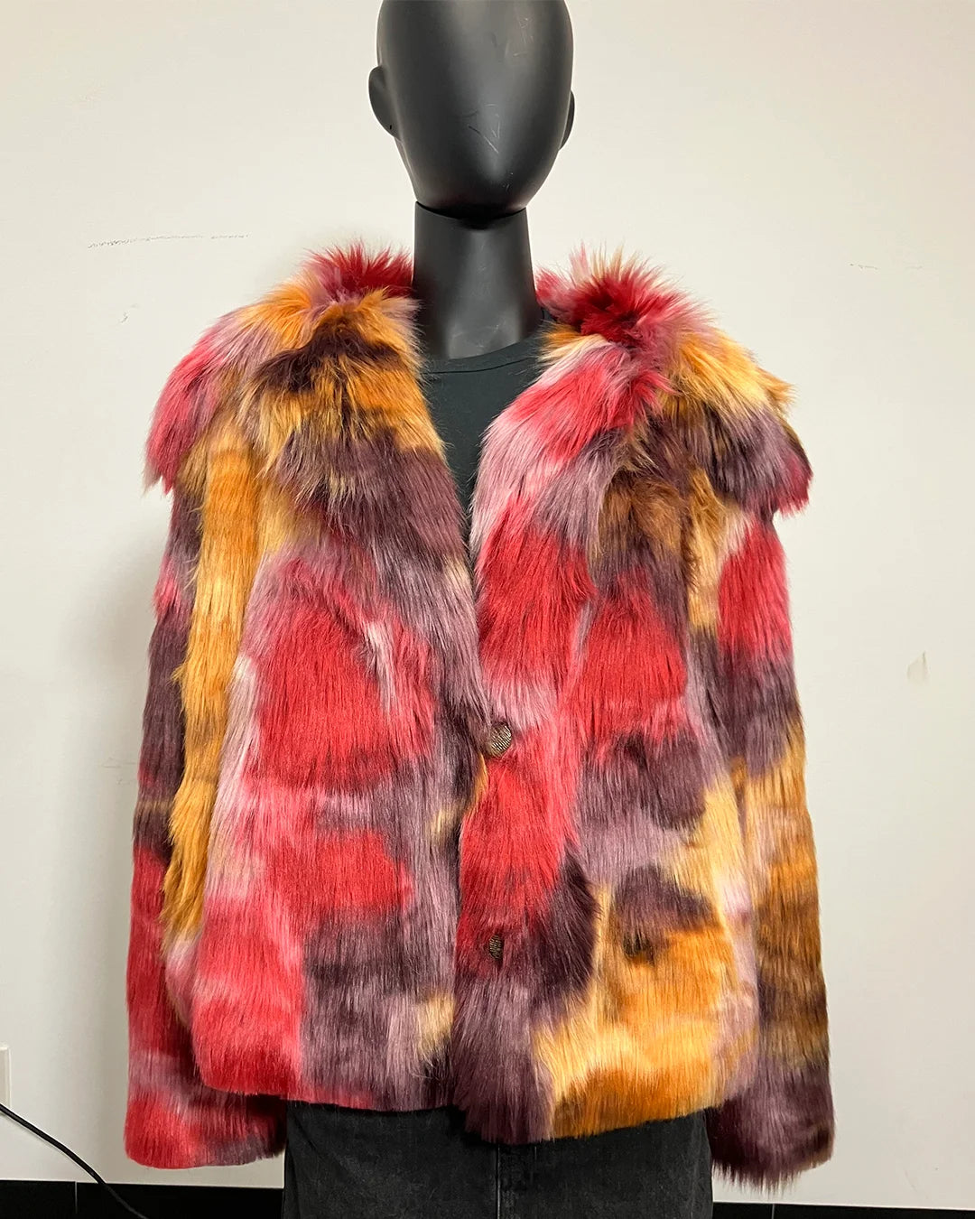 A multicolor faux fur coat with red, orange, yellow, and purple tones, displayed on a black mannequin in front of  a plain off-white wall.