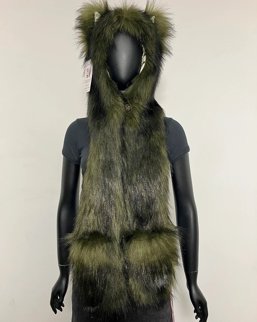A long, shaggy faux-fur hooded scarf or vest in olive green and black, displayed on a black mannequin against a plain off-white wall.