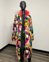 A long, open-front faux fur coat with a multicolor pattern of pink, yellow, green, white, and black, featuring a bright pink collar, displayed on a black mannequin in an indoor room setting.