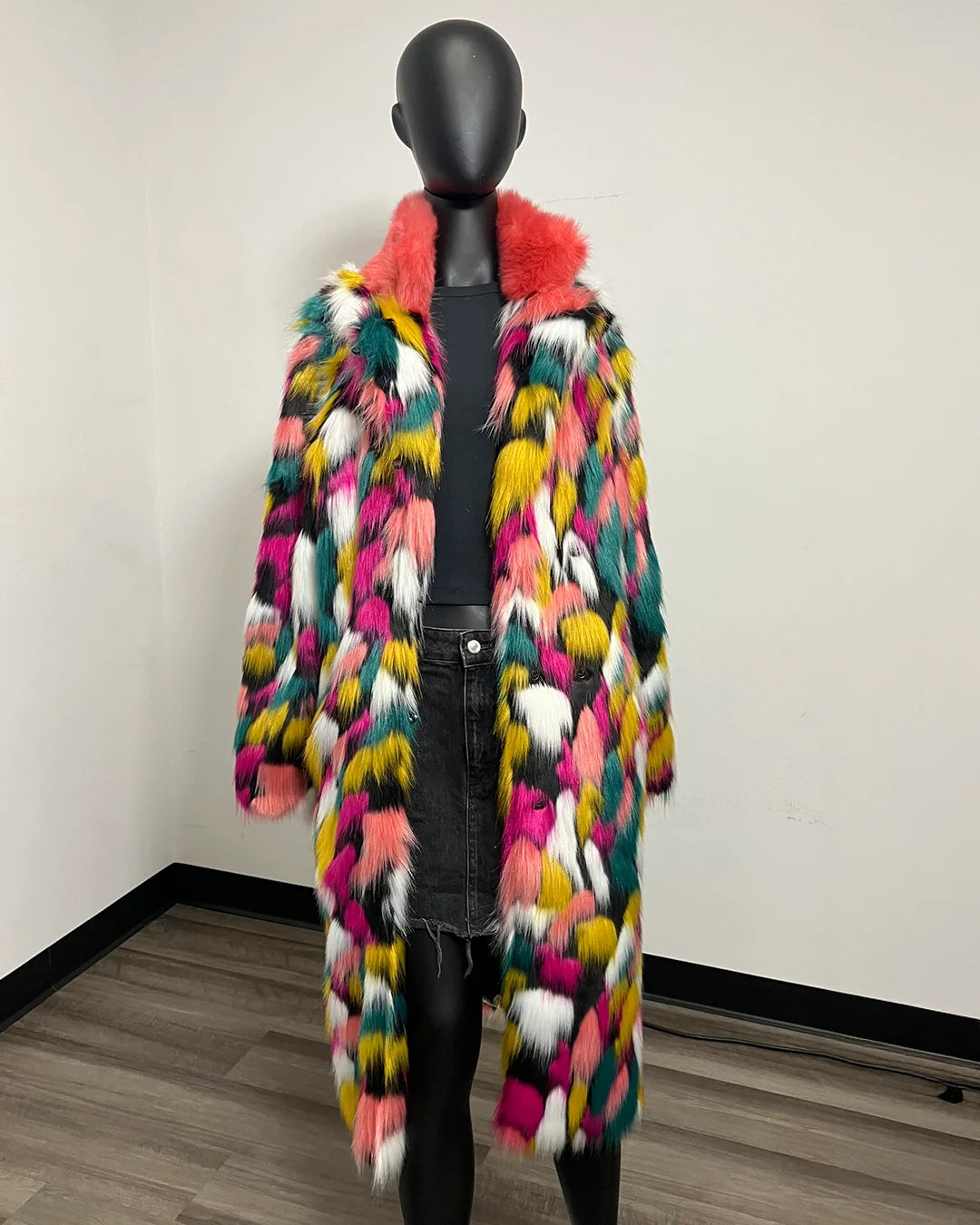 A long, open-front faux fur coat with a multicolor pattern of pink, yellow, green, white, and black, featuring a bright pink collar, displayed on a black mannequin in an indoor room setting.