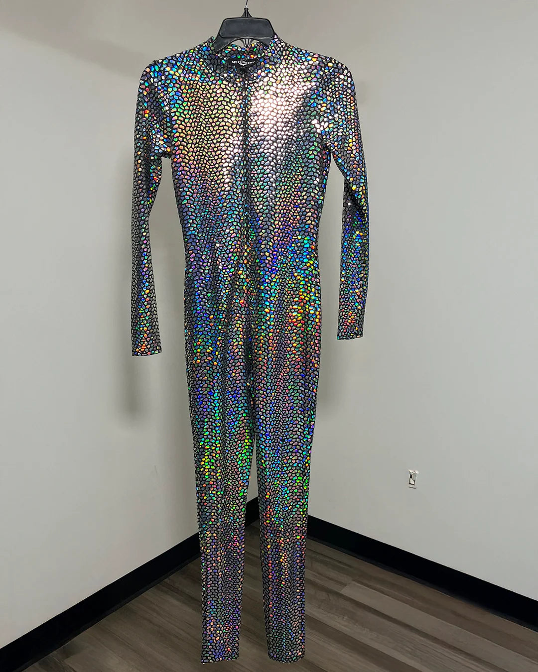 A long-sleeve, full-length one-piece jumpsuit covered in iridescent holographic snakeskin pattern foil print, hanging on a black hanger against a plain indoor wall.