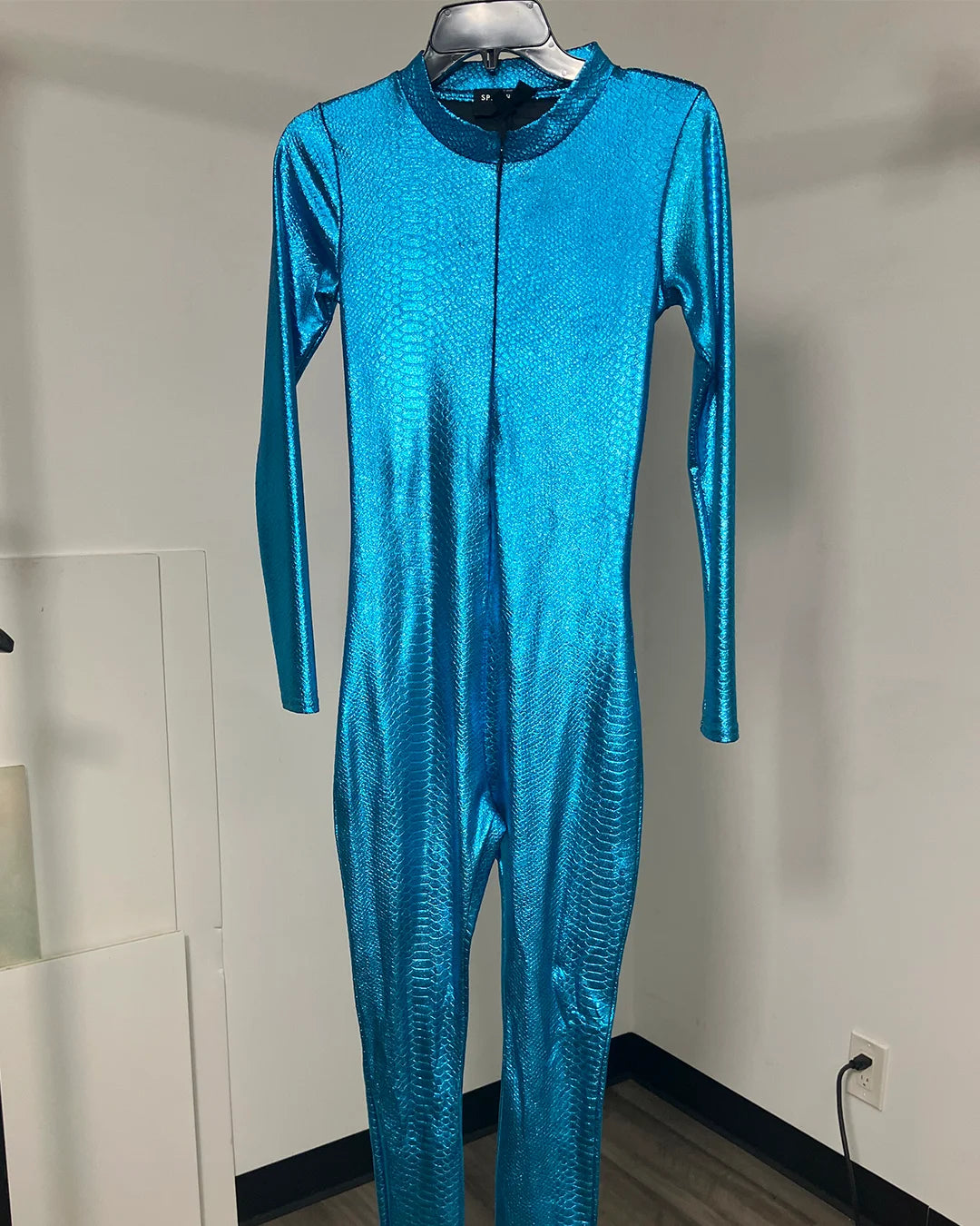 A shiny metallic blue one-piece jumpsuit or bodysuit displayed on a hanger against a plain indoor wall.