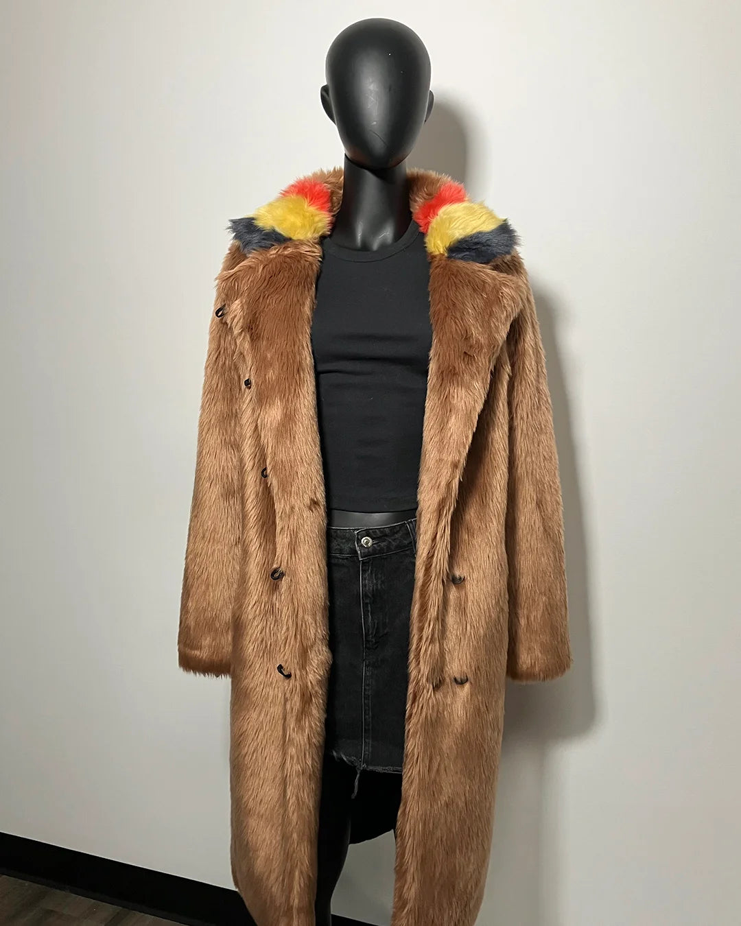 Mannequin wearing a brown faux fur coat with colorful hood trim against a white wall.