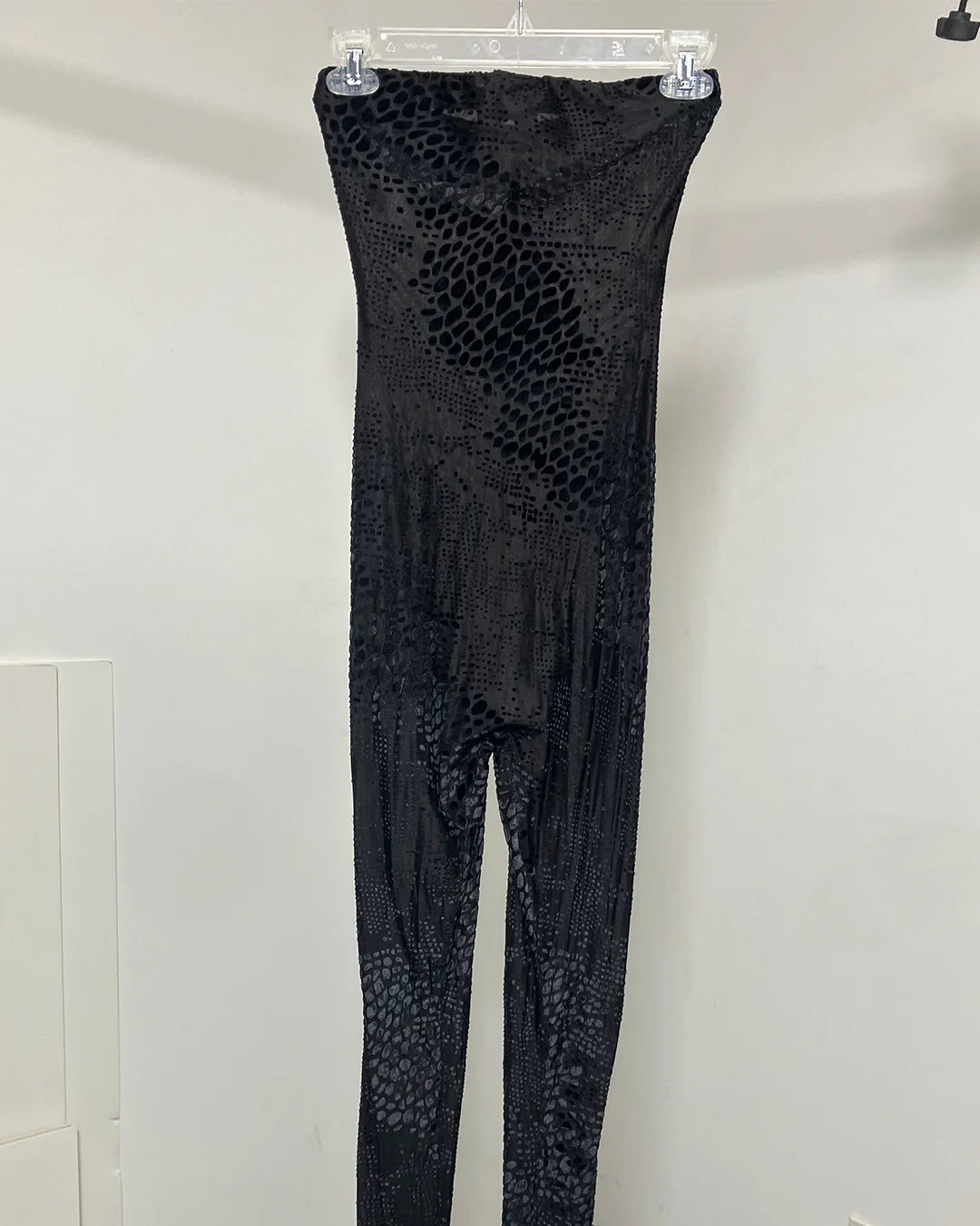 Black snakeskin-patterned strapless jumpsuit hanging on a clear plastic hanger against a plain white wall.