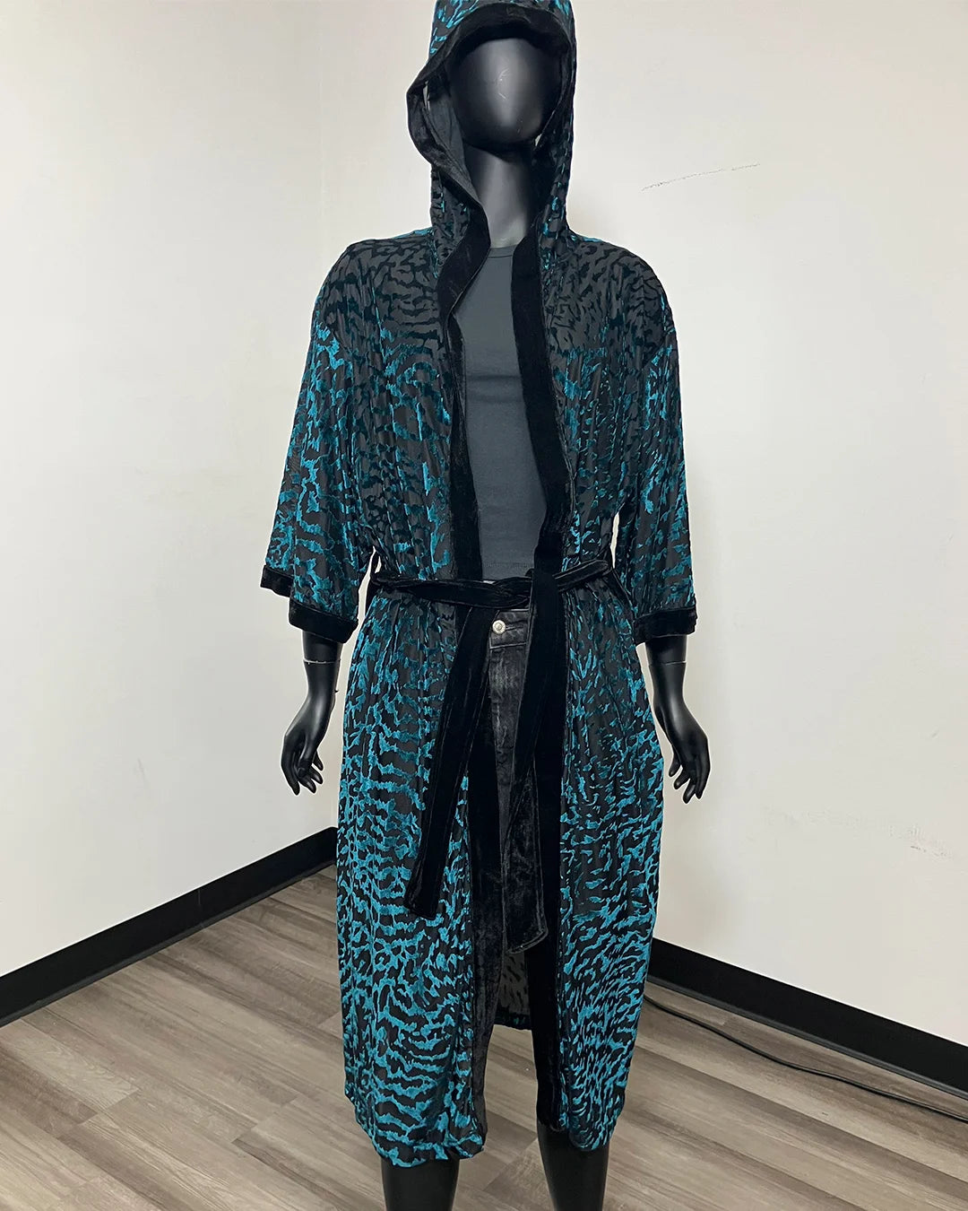 A hooded robe with a teal and black animal print pattern, displayed on a black mannequin in an indoor room with a white wall and wood floor.
