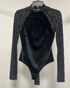 A black long-sleeve bodysuit with a leopard-print mesh pattern on the sleeves and shoulders, hanging on a black hanger against a plain off-white wall.