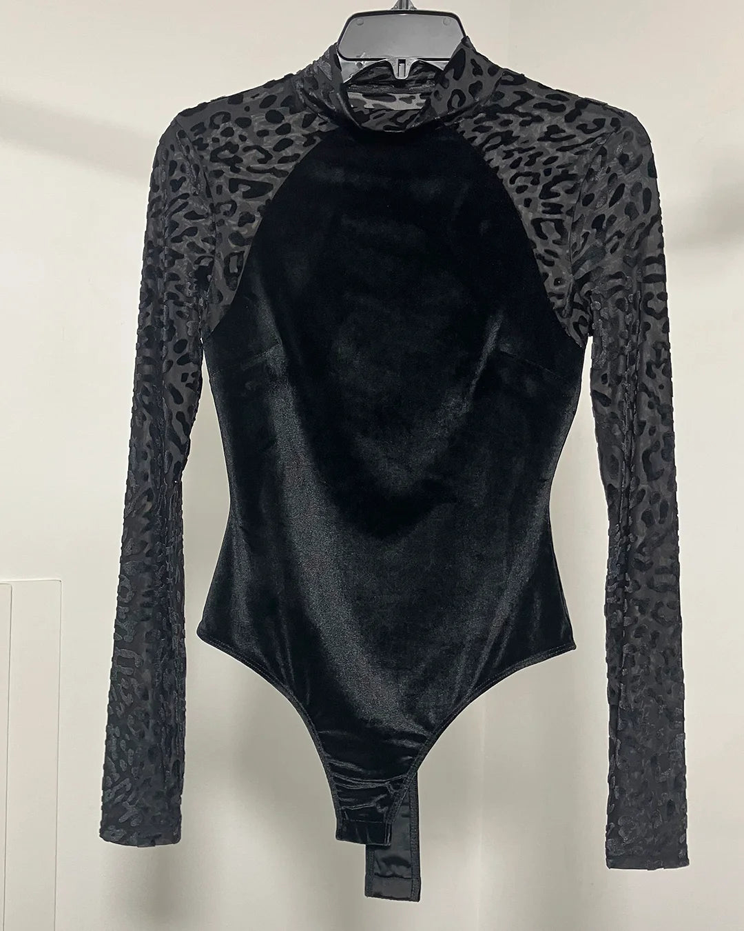 A black long-sleeve bodysuit with a leopard-print mesh pattern on the sleeves and shoulders, hanging on a black hanger against a plain off-white wall.