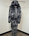 A one-piece jumpsuit with a a zip-up front and hood with ears, featuring a black and gray marbled tie-dye pattern, hanging on a metal clothes rack against a plain wall.