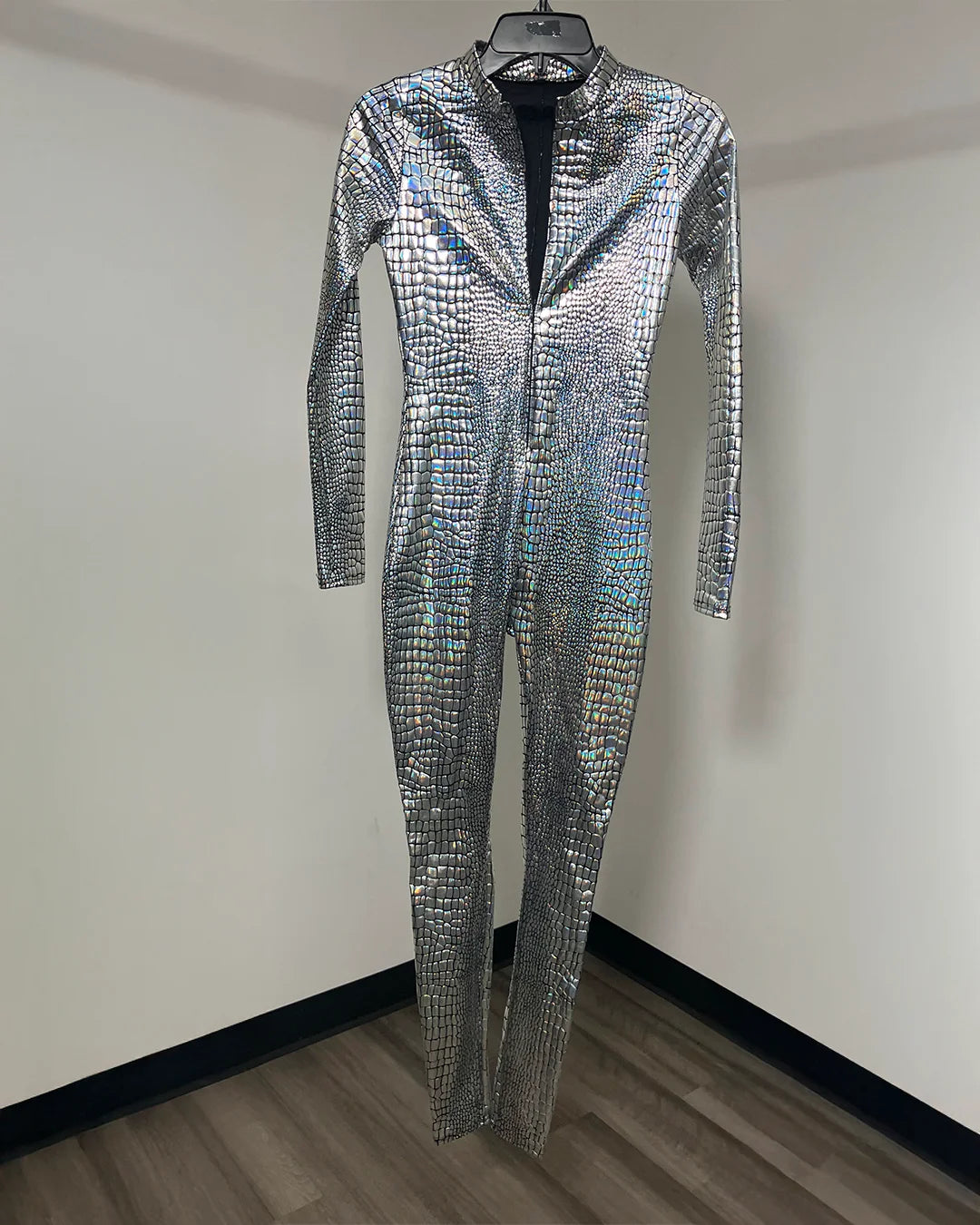 A long-sleeve, full-length metallic iridescent silver jumpsuit with a textured, reptile-like scale pattern, hanging on a black hanger against a plain indoor wall.