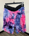Fuzzy tie-dye shorts in pink, purple, and blue hanging from a black hanger against an indoor wall.