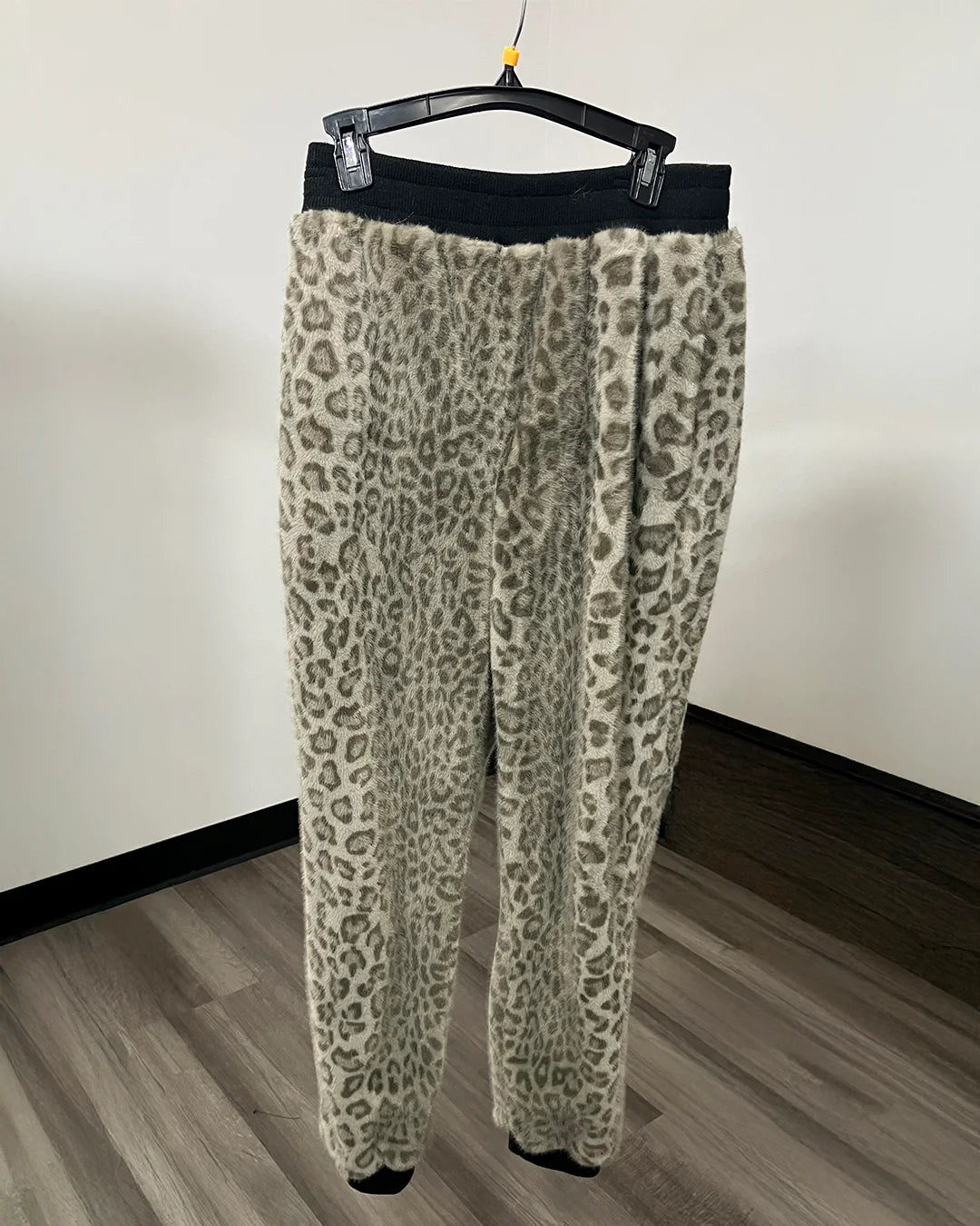Leopard print fleece pants hanging on a black hanger against a plain wall and wood floor.