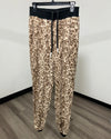 Leopard print fleece jogger pants with a black elastic waistband and drawstring, hanging on a black clothes hanger against a plain white wall and wood-look floor.