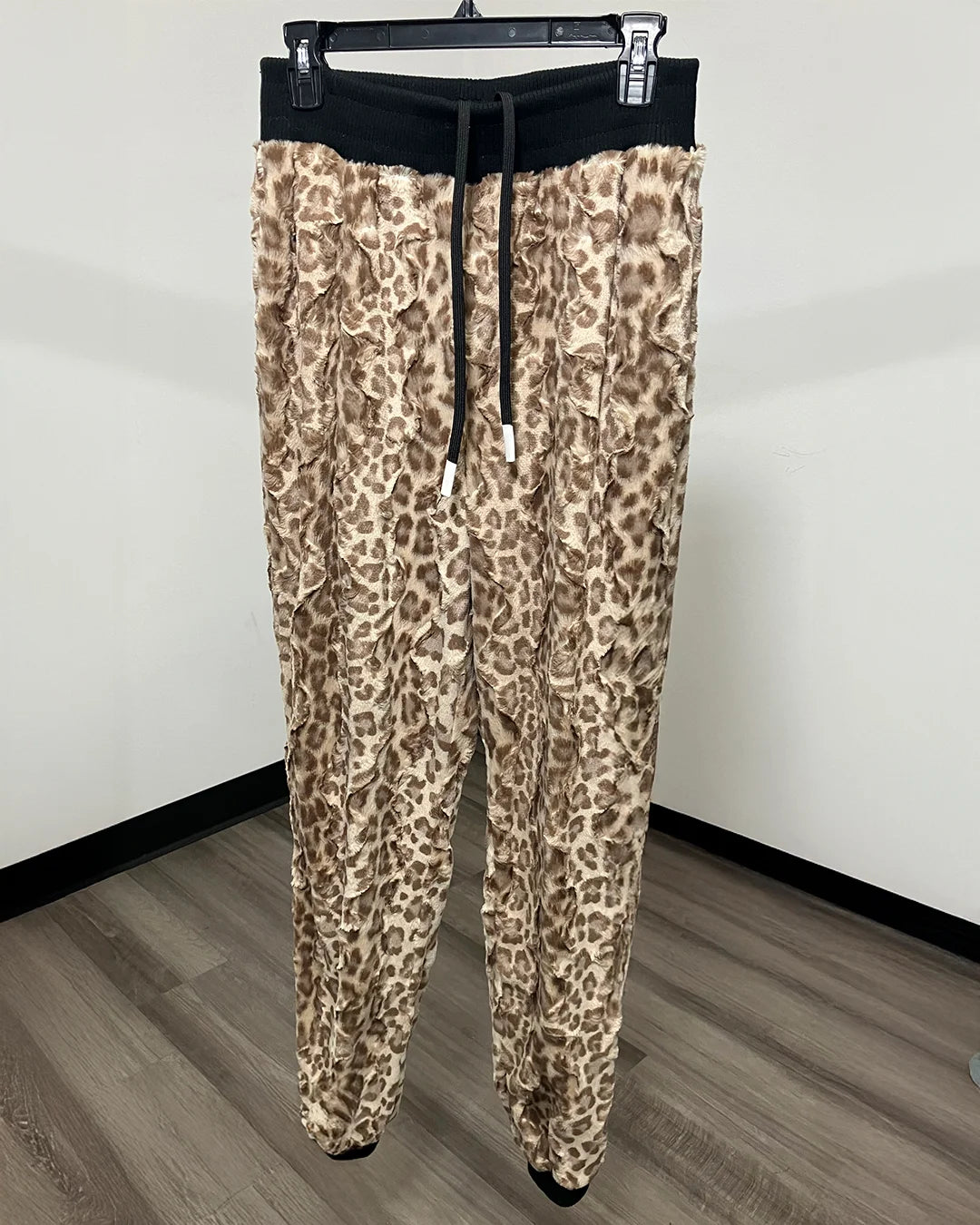 Leopard print fleece jogger pants with a black elastic waistband and drawstring, hanging on a black clothes hanger against a plain white wall and wood-look floor.