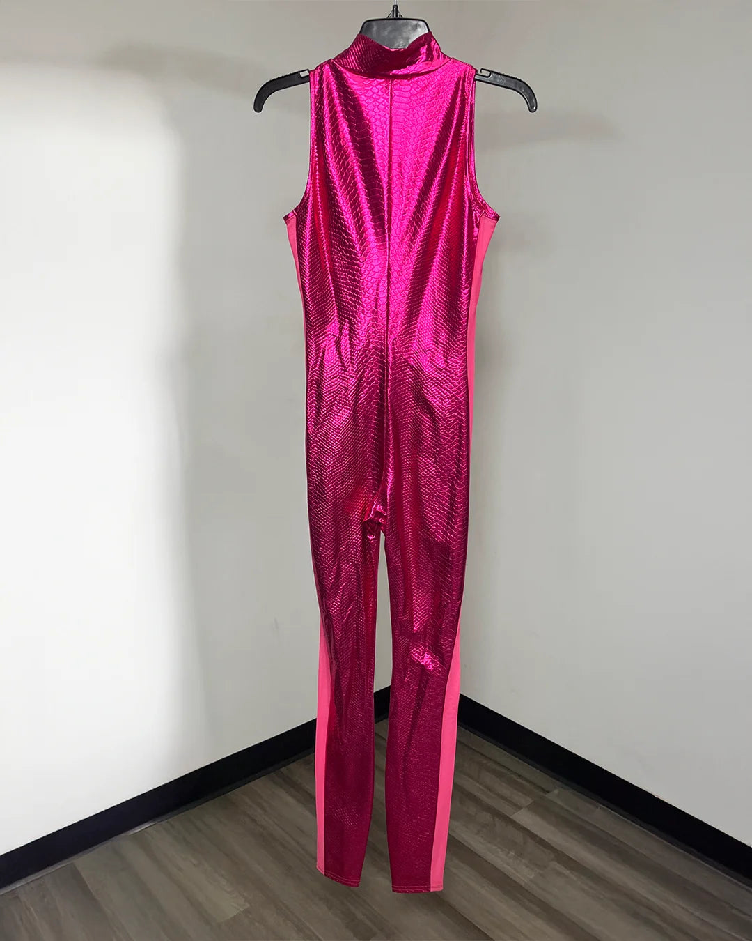 A sleeveless, high-neck, full-length jumpsuit in a metallic hot pink color with a subtle textured pattern, hanging on a black hanger against a plain white wall.