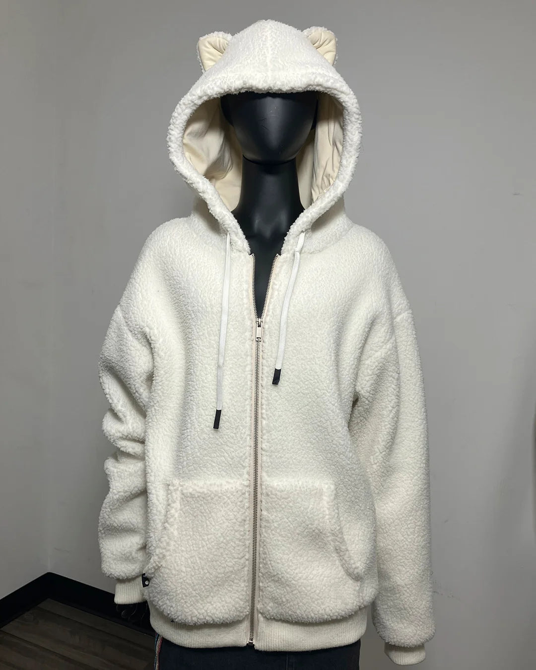 An off-white sherpa fleece zip-up hoodie with a hood featuring bear ears and a cream-colored lining, displayed on a black mannequin against a plain gray wall.