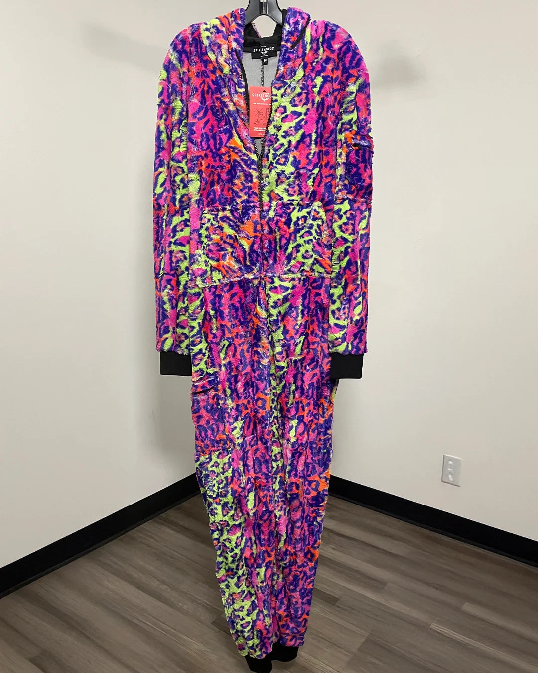 A zip-up hooded onesie featuring a vibrant multicolor abstract pattern in purple, pink, orange, and green. The onesie is displayed on a hanger against a plain white wall with a wood-look floor.