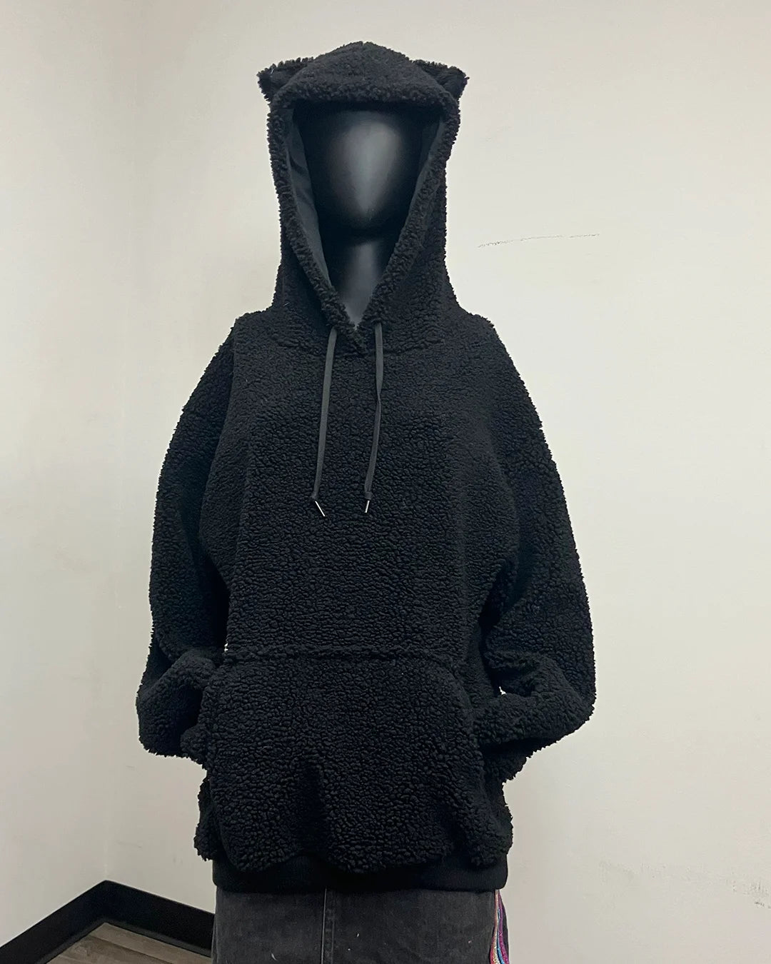 A black sherpa fleece hoodie with a plush teddy texture, displayed on a black mannequin. The hoodie features a hood with cat-like ears, dark gray drawstrings, and a front kangaroo pocket.