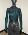 A long-sleeve mock neck crop top with a black and teal abstract pattern, displayed on a black mannequin torso wearing a gray denim skirt.