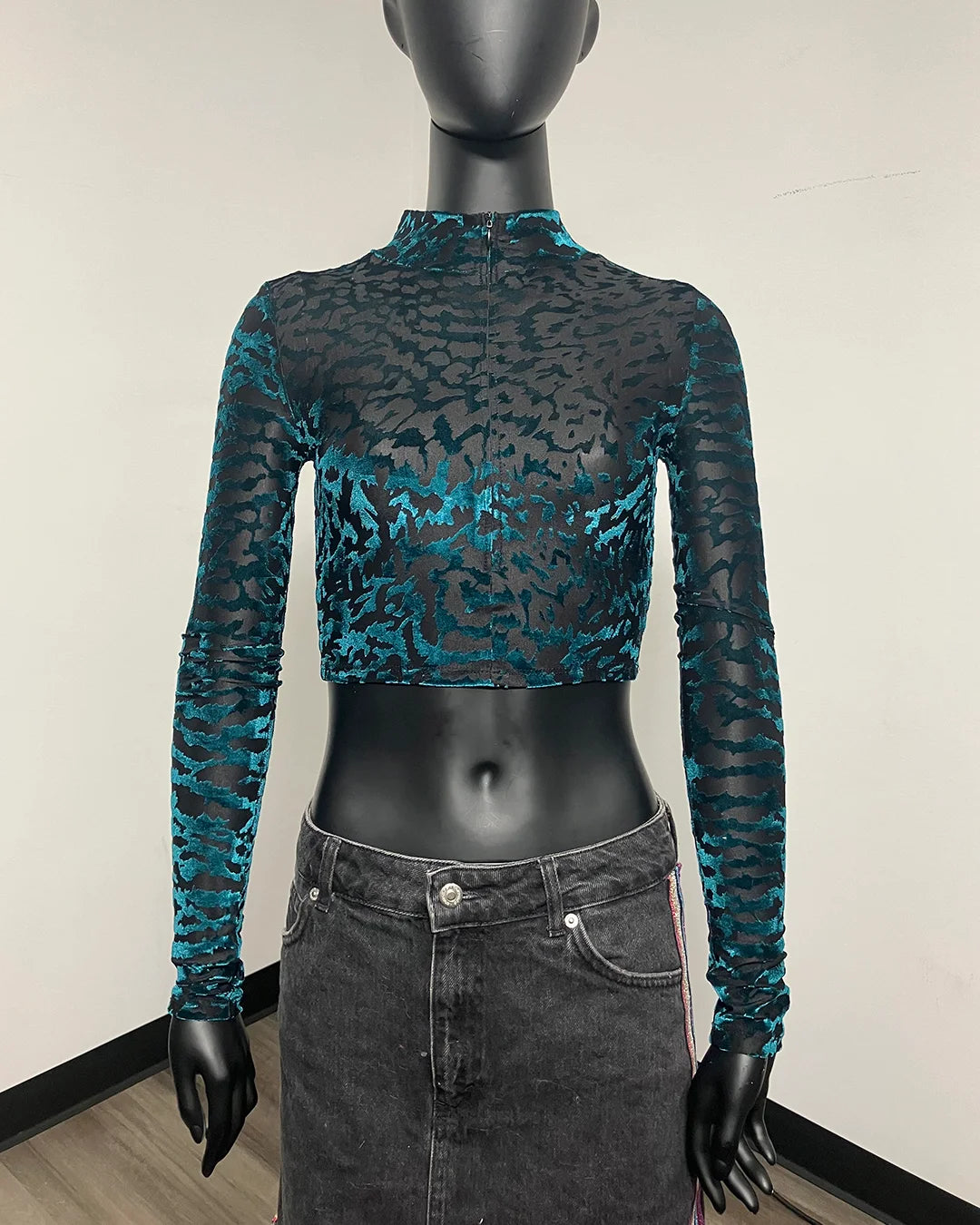 A long-sleeve mock neck crop top with a black and teal abstract pattern, displayed on a black mannequin torso wearing a gray denim skirt.