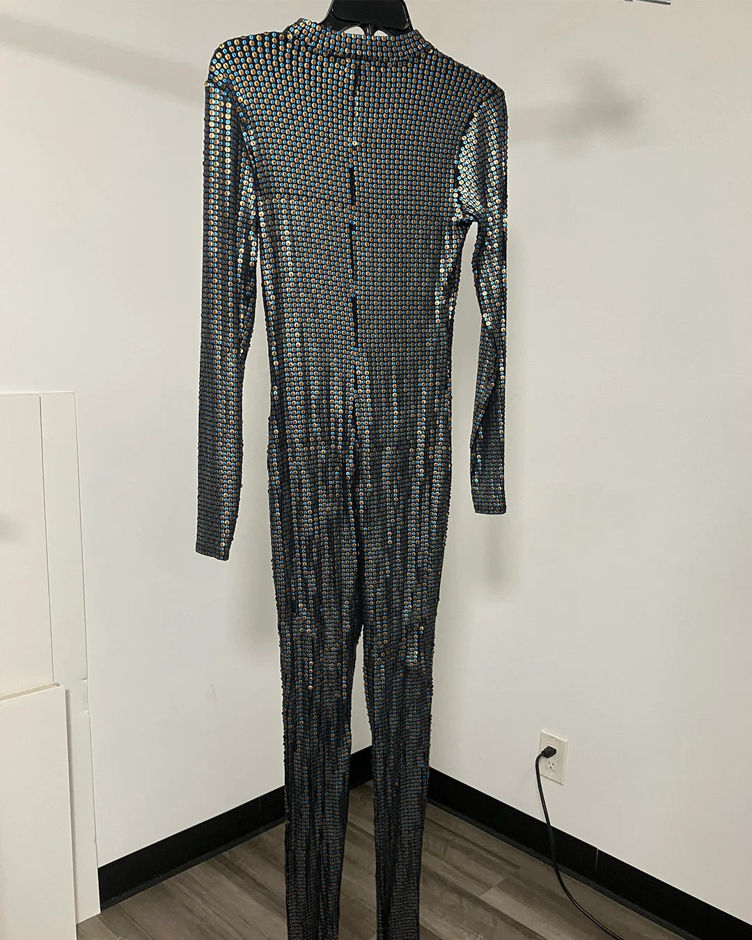 A long-sleeve, full-length jumpsuit covered in shimmering silver sequins, displayed on a black hanger against a plain white wall.