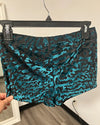 A pair of women's shorts with a black and teal abstract pattern, hanging on a black hanger with a hand holding the left hem.