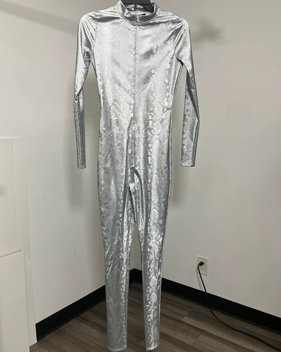 A full-body metallic silver jumpsuit with a textured, crinkled pattern, hanging on a black hanger against a plain white wall.