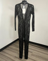 A black sheer lace long-sleeve jumpsuit with a front zipper, hanging on a black hanger against a plain indoor wall with wood-look flooring.