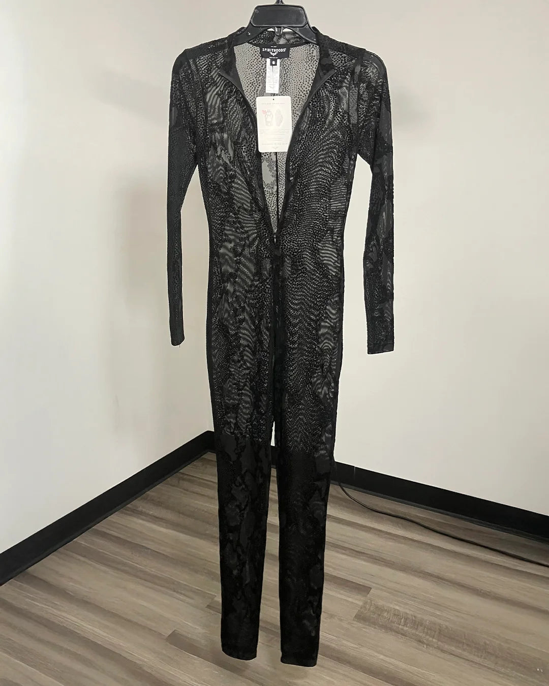A black sheer lace long-sleeve jumpsuit with a front zipper, hanging on a black hanger against a plain indoor wall with wood-look flooring.